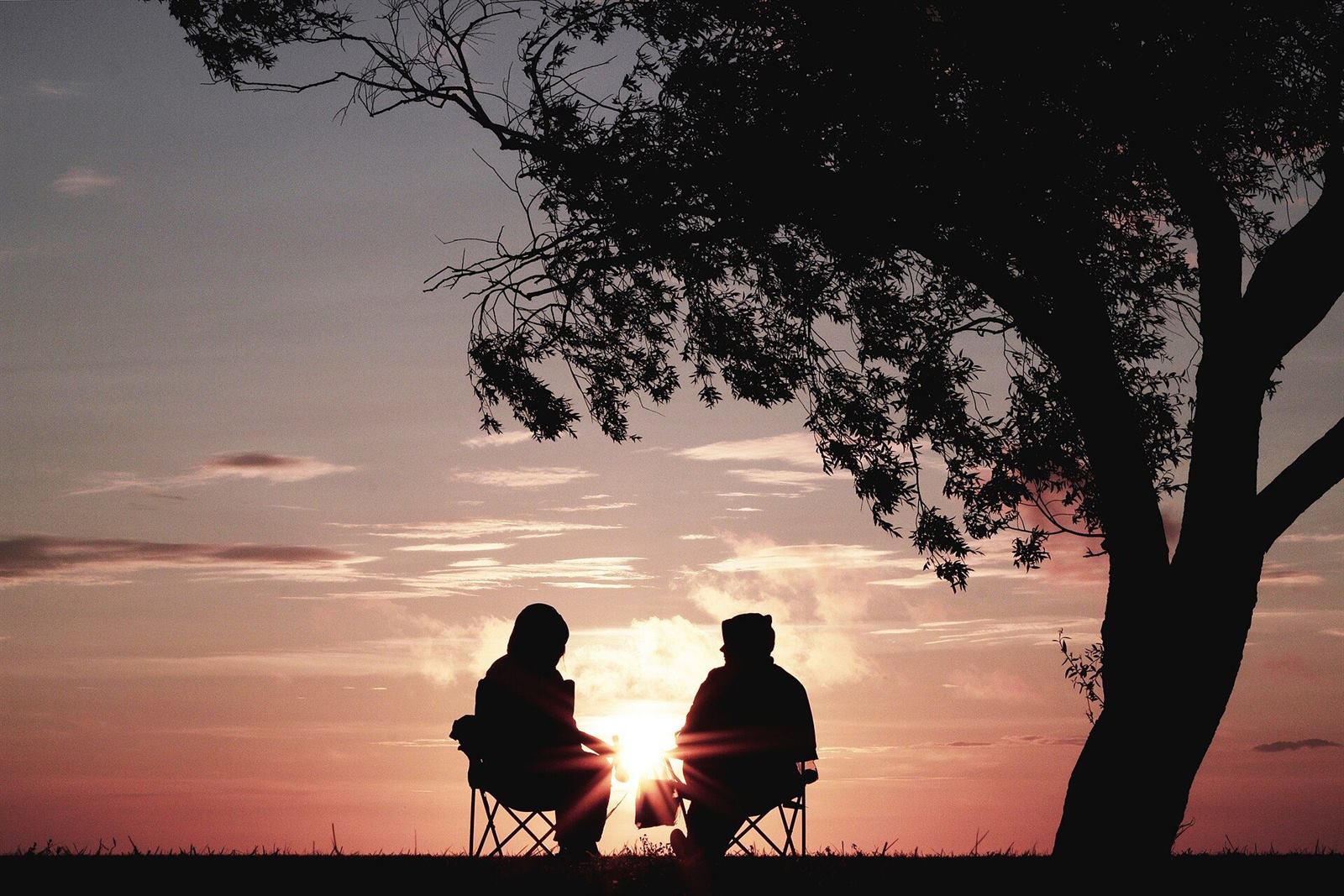 Two people in camp chairs watching a pink sunset under a tree