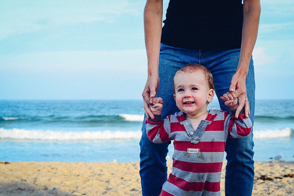 A toddler taking first steps on the beach, holding a parent's hands